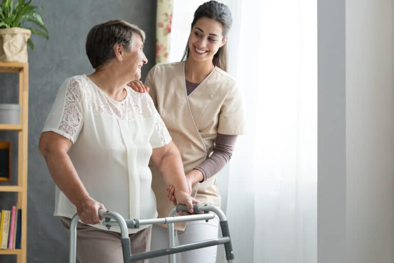 Caregiver in a beige tunic helping an older woman stand with a walker beside a bright window at home